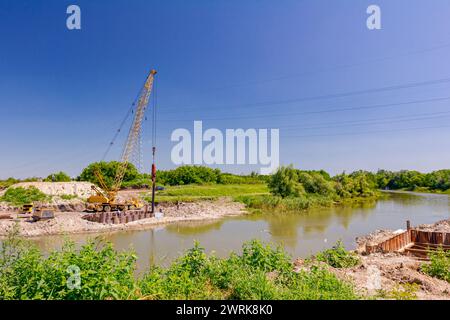 Across the water is yellow obsolete mobile crane with pneumatic hammer. Installed metal piles along river bank built in for bridge foundation at the c Stock Photo