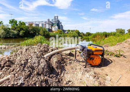 Orange motor pump with long hose for filling tank with water placed on coast, bridge construction is in progress, across the river are big grain silos Stock Photo