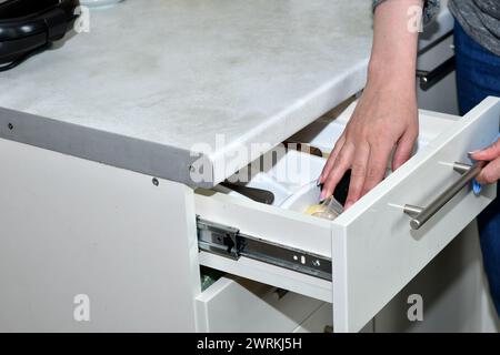 With one hand, the hostess opened a drawer in the kitchen furniture, and with the other she tries to take cutlery. Stock Photo