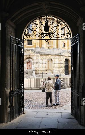 View through Bodleian Library wrought iron gate arch with senior couple standing in Radcliffe Square next to Radcliffe Camera, University of Oxford UK Stock Photo