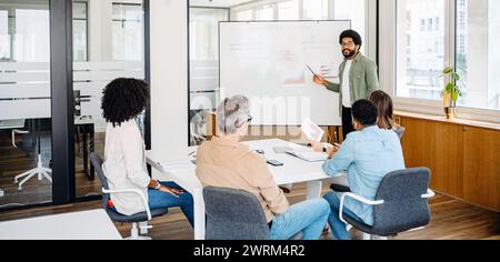 A diverse and dynamic team gathers in a modern office space, where a young professional takes the lead in presenting strategic insights on a whiteboard. The ideas and strategies exchanging Stock Photo