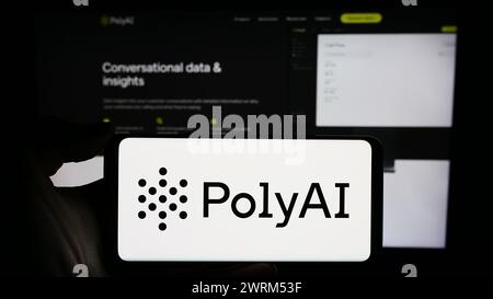 Person holding cellphone with logo of conversational artificial intelligence company PolyAI Ltd. in front of webpage. Focus on phone display. Stock Photo