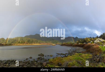 A rainbow is seen over a body of water with a house in the background. The sky is cloudy and the water is calm Stock Photo