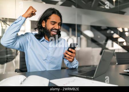 Excited indian guy manager looking at smartphone screen, gesturing Stock Photo