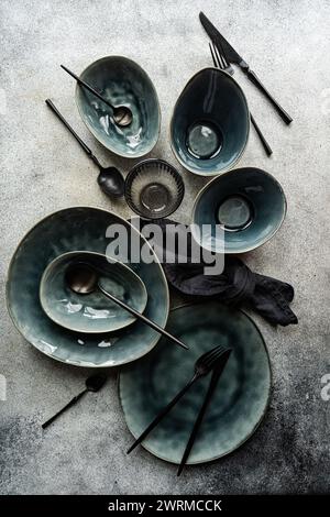 Top view of a luxurious table setting featuring emerald green ceramic plates, bowls, and dark cutlery on a textured grey surface. Stock Photo
