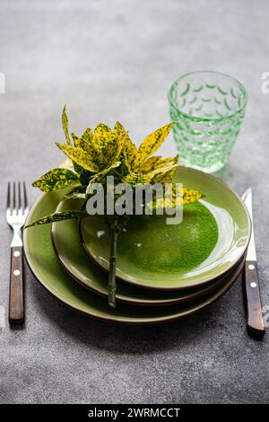 Elegant table setting featuring bright green ceramic plates, a decorative plant, cutlery, and a patterned glass on a textured surface. Stock Photo