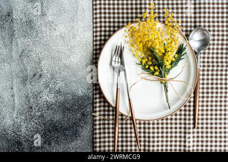 From above of white plate with a border detail, elegantly set with copper-toned cutlery and adorned with a cheerful mimosa flower arrangement, atop a Stock Photo