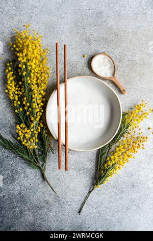 Top view of elegant minimalist table setting, with a ceramic plate and chopsticks, accented by the bright yellow of mimosa flowers against a textured Stock Photo