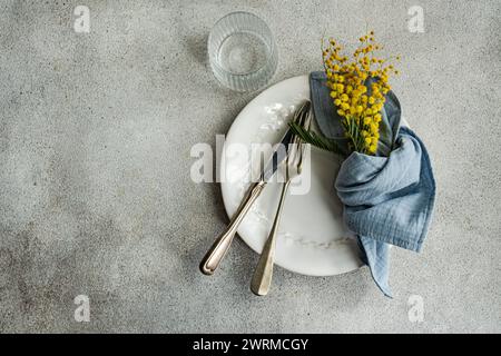 Elegant dining setup featuring a white ornate plate, vintage silverware, a blue linen napkin, and a bright yellow mimosa bouquet on a textured backdro Stock Photo
