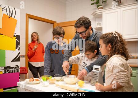A family enjoys a cooking session together in a well-lit kitchen, with the father guiding the children in preparing a meal. Stock Photo