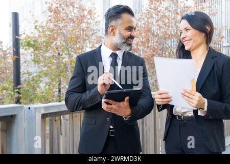 Professional man and woman discussing work with papers and a digital tablet, smiling in an outdoor urban setting. Stock Photo