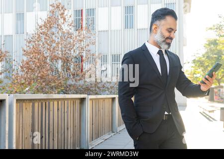 A suited man with a beard smiles while using his phone outside an office building. Stock Photo