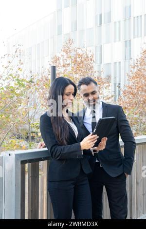 A professional man and woman engage in a work discussion while reviewing data on a tablet computer outdoors in an urban setting. Stock Photo