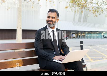 A professional man in a suit works on his laptop while seated on a public bench, smiling and enjoying the outdoor setting. Stock Photo