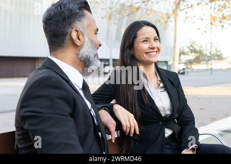 A man and woman in business attire share a light moment on a sunny day, reflecting a positive work environment. Stock Photo