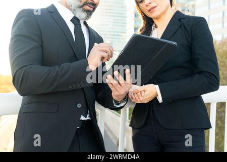A professional man and woman in business attire stand outside, reviewing data on a digital tablet. Stock Photo
