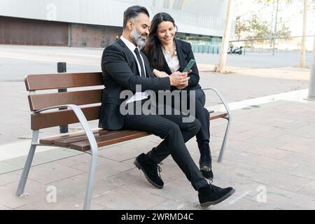 A man and woman in business attire sit close together on a park bench, smiling as they look at a smartphone. Stock Photo