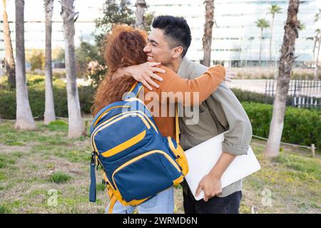 An affectionate hug between a woman and a man on a college campus, with the woman carrying a blue and yellow backpack and the man holding a white lapt Stock Photo