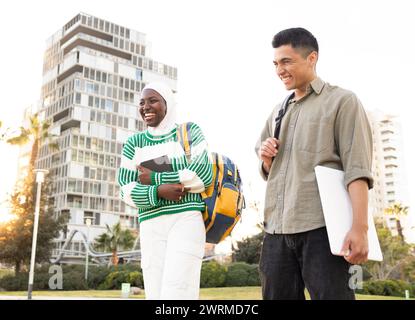 A joyful African American woman and latin man engaging in a friendly conversation, sharing a laugh while holding their study materials on a sunny day Stock Photo