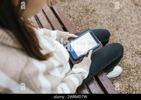 Over the shoulder shot of a young female engrossed in reading an ebook on her digital tablet outdoors on a wooden bench. Stock Photo