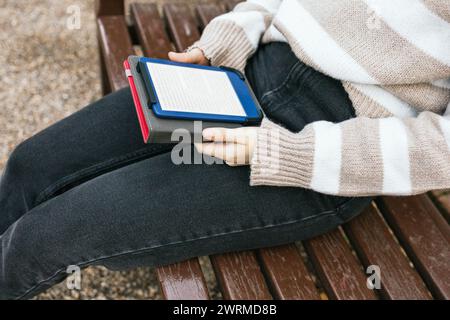 Close-up of an individual's lap while sitting on a bench with an e-book reader in hand, set against a casual outdoor backdrop. Stock Photo