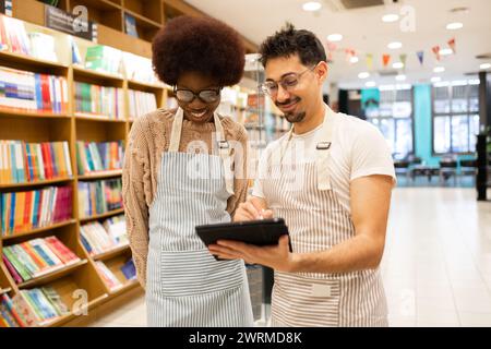 Two cheerful bookstore workers in aprons sharing a tablet and smiling while standing in a well-organized bookshop Stock Photo