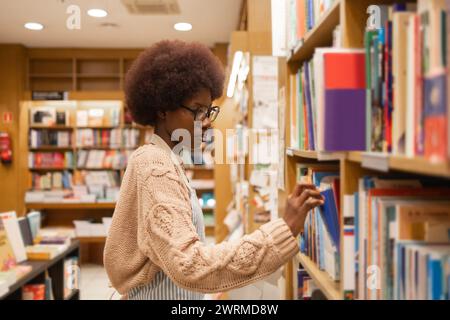 Side view of young black African American woman with glasses and a cozy sweater is thoughtfully selecting books from the shelves in a well-lit library Stock Photo