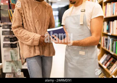 A close-up view of two cropped unrecognizable individuals in a cozy library, one holding and reading a book while engaging in a discussion Stock Photo