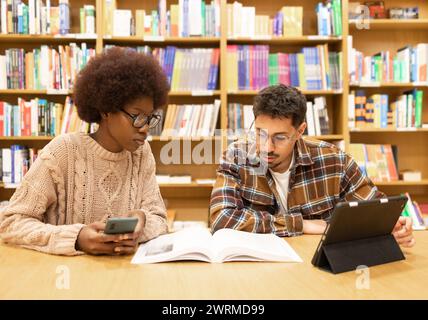 Two students engaged in studying at a library table with books and digital tablets, amidst rows of bookshelves Stock Photo