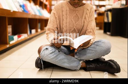 A cropped unrecognizable person sits on the floor of a library, deeply focused on reading a book, with shelves of books in the background Stock Photo