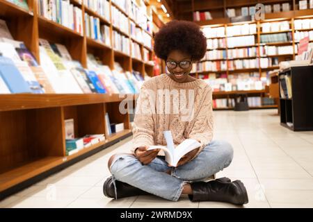 A joyful African American woman sits cross-legged on the library floor, immersed in reading a book among towering bookshelves Stock Photo