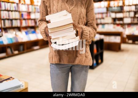 Close-up image of an unrecognizable individual in a cozy sweater holding a large pile of books, standing in the aisle of a well-stocked library Stock Photo