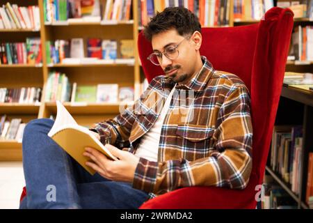 A young adult man engrossed in reading a book while sitting in a red armchair, surrounded by bookshelves in a library. Stock Photo