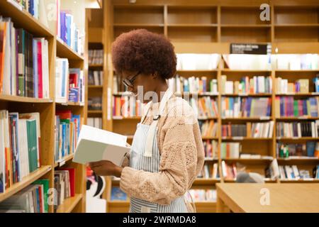Side view of young African American woman with a curly afro is engrossed in reading a book among library shelves filled with literature Stock Photo