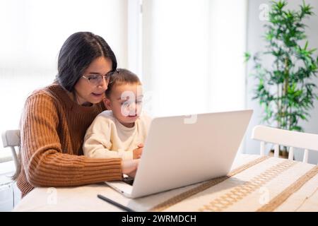 A heartwarming moment as a positive mother embraces her son while they both engage with content on a laptop at home Stock Photo