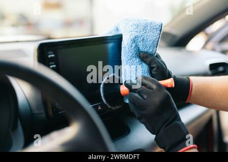 A person wearing black gloves gently cleans a car's infotainment screen using a soft microfiber towel and detailing brush Stock Photo