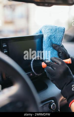 Close-up view of a person's hands cleaning a car's interior using a blue microfiber towel and a detailing brush Stock Photo
