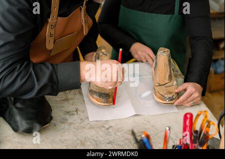 A cropped unrecognizable shoemaker carefully traces a pattern for a shoe sole on white paper in a well-equipped workshop. Stock Photo