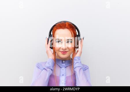 Captured against a white backdrop stands a businesswoman in her vibrant red hair and a professional lilac blouse using wireless headphones looking awa Stock Photo