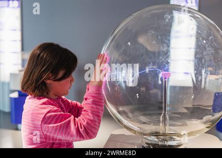 A curious girl in a pink striped shirt engages with a plasma globe exhibit, fascinated by the physics demonstration at a science museum. Stock Photo