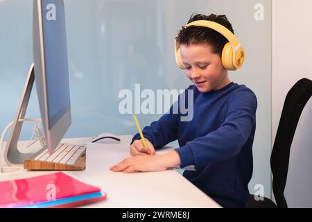 A dedicated child wearing headphones while writing notes at a desk with a computer, illustrating a modern multi-tasking study environment Stock Photo