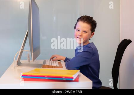 A happy child with a bright smile sits at a desk, using a computer with colorful notebooks stacked beside him, ready for learning Stock Photo