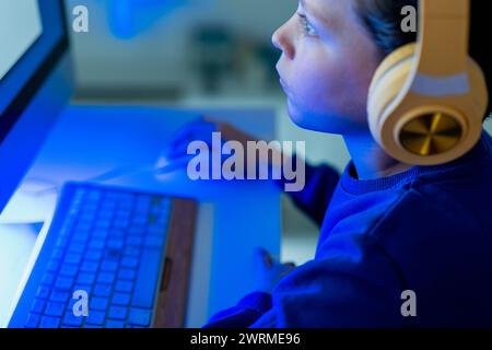 A child is deeply focused on the glowing computer screen, with headphones on, in a darkened room illuminated by the screen's light Stock Photo