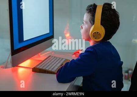A child is intently using a computer, illuminated by the warm backlight of the monitor, with headphones on Stock Photo