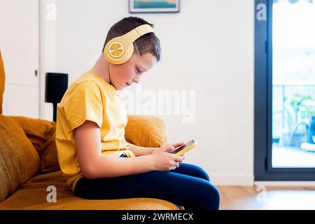 A focused boy sits on a sofa, wearing yellow headphones and using a smartphone, with a cozy home interior in the background Stock Photo