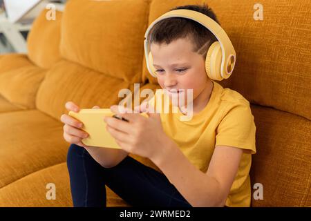 A focused boy wearing yellow headphones plays games on a smartphone while sitting on a sofa Stock Photo
