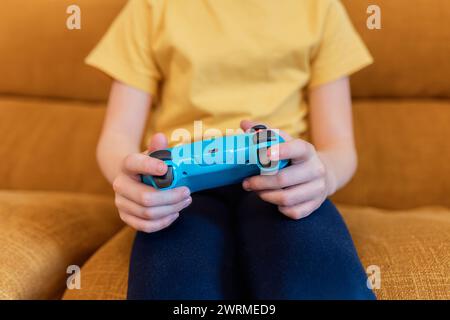 Close-up of a child's hands holding a vibrant blue game controller, focused on gaming while sitting on a cozy sofa Stock Photo