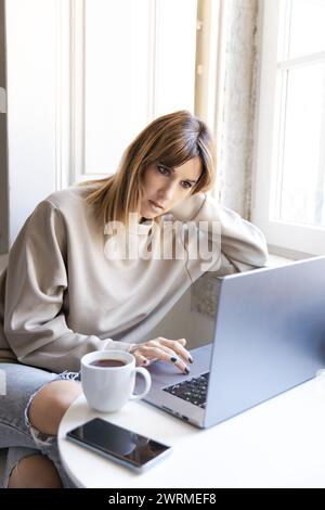A thoughtful woman works on her laptop with coffee and smartphone nearby Stock Photo
