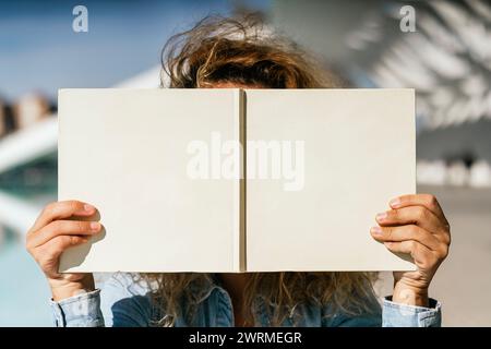 In a striking outdoor setting, anonymous person with hands that have been surgically adapted due to no thumbs holds an open book, covering their face Stock Photo
