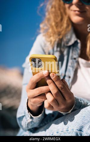 A close-up view captures an woman with sunglasses, holding a smartphone with a yellow case, focusing on the hands interacting with the device under a Stock Photo